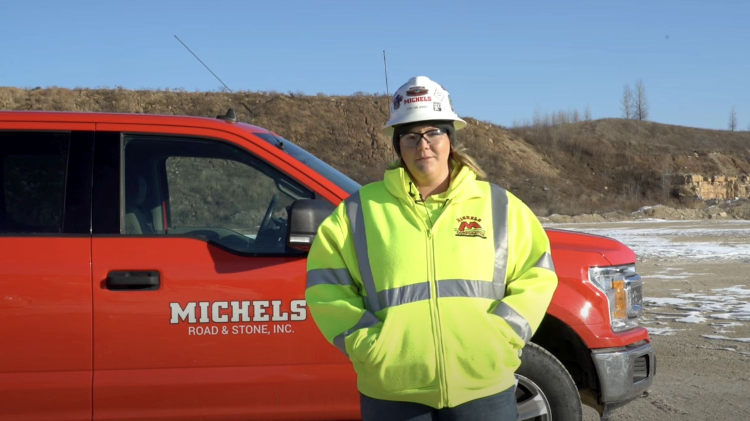 A woman wearing a vi-vis jacket and hardhat stands next to a red Michels Road & Stone Truck.