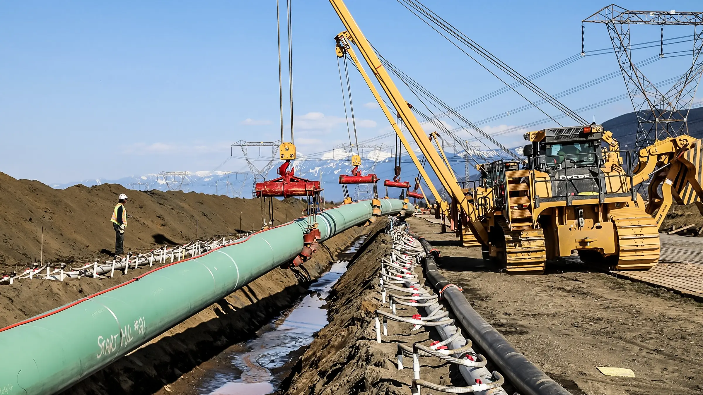 Pipelayers lower a large diameter pipeline into a trench in a rural Canadian environment.