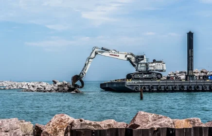 An excavator works on a barge to lift shoreline rocks.