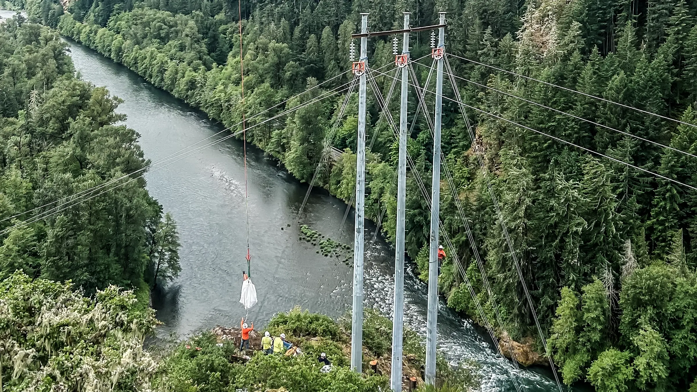 A Helicopter assists a crew on a tall transmission tower near a river.