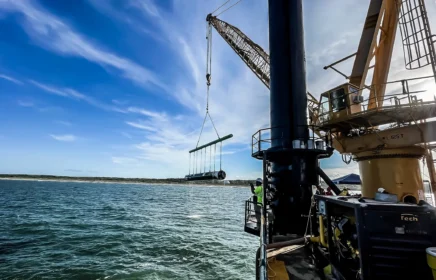 A crane aboard a barge lifts a piece of pipe over water.