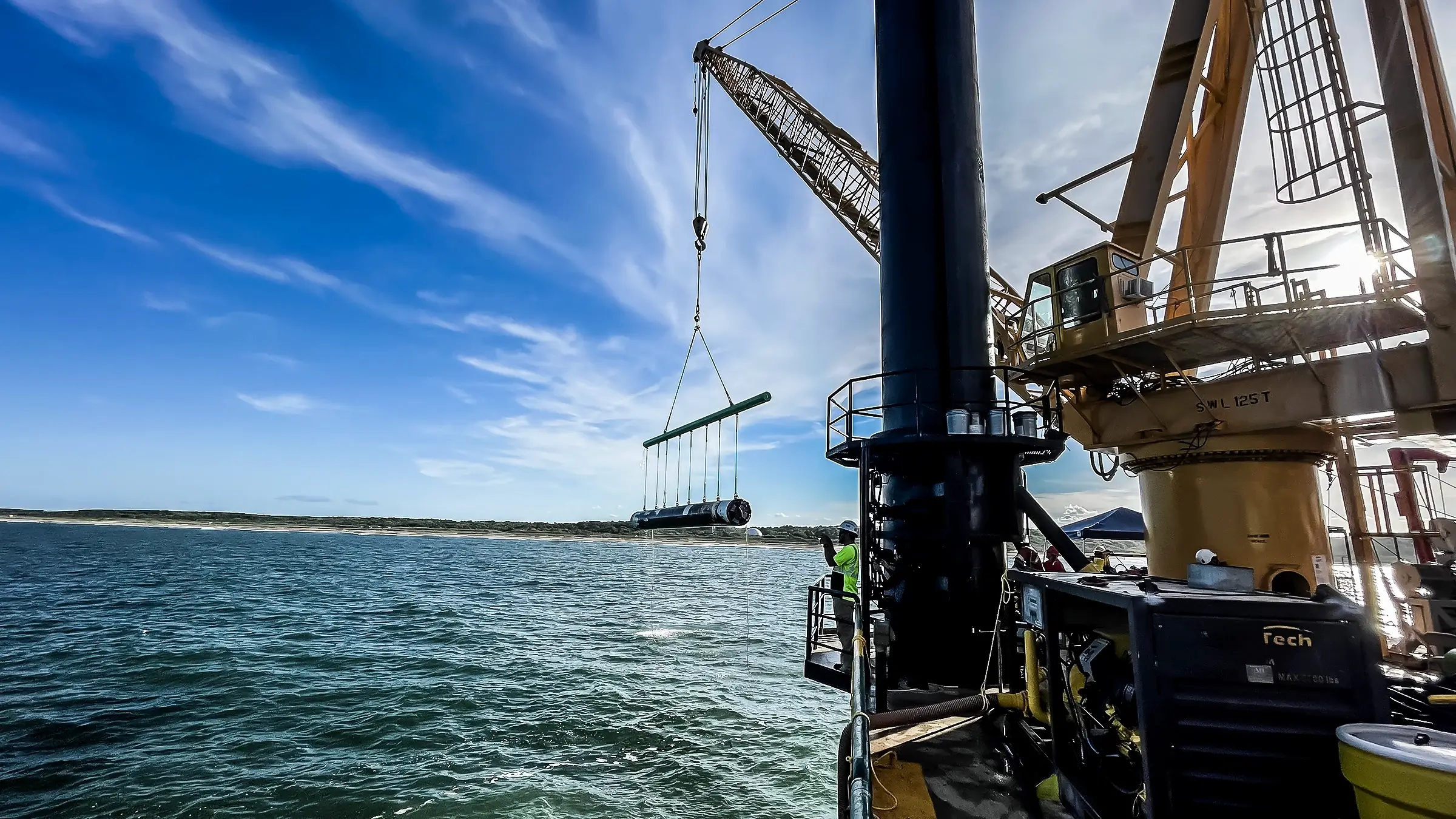 A crane aboard a barge lifts a piece of pipe over water.